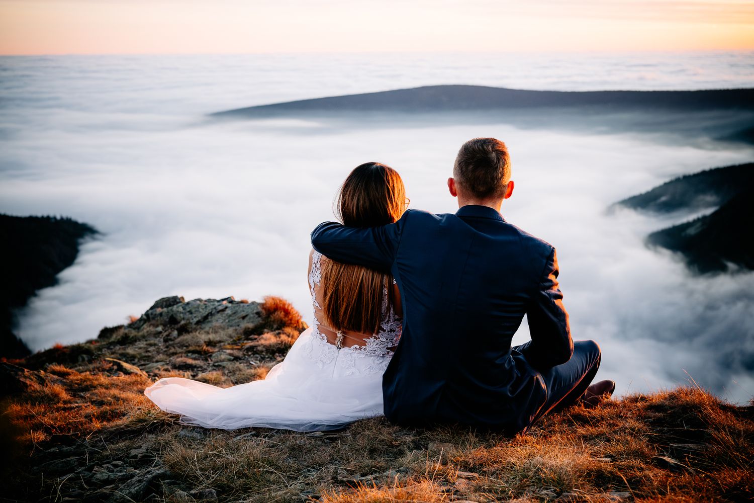 Couple sits together on mountaintop overlooking sea of clouds at sunset.