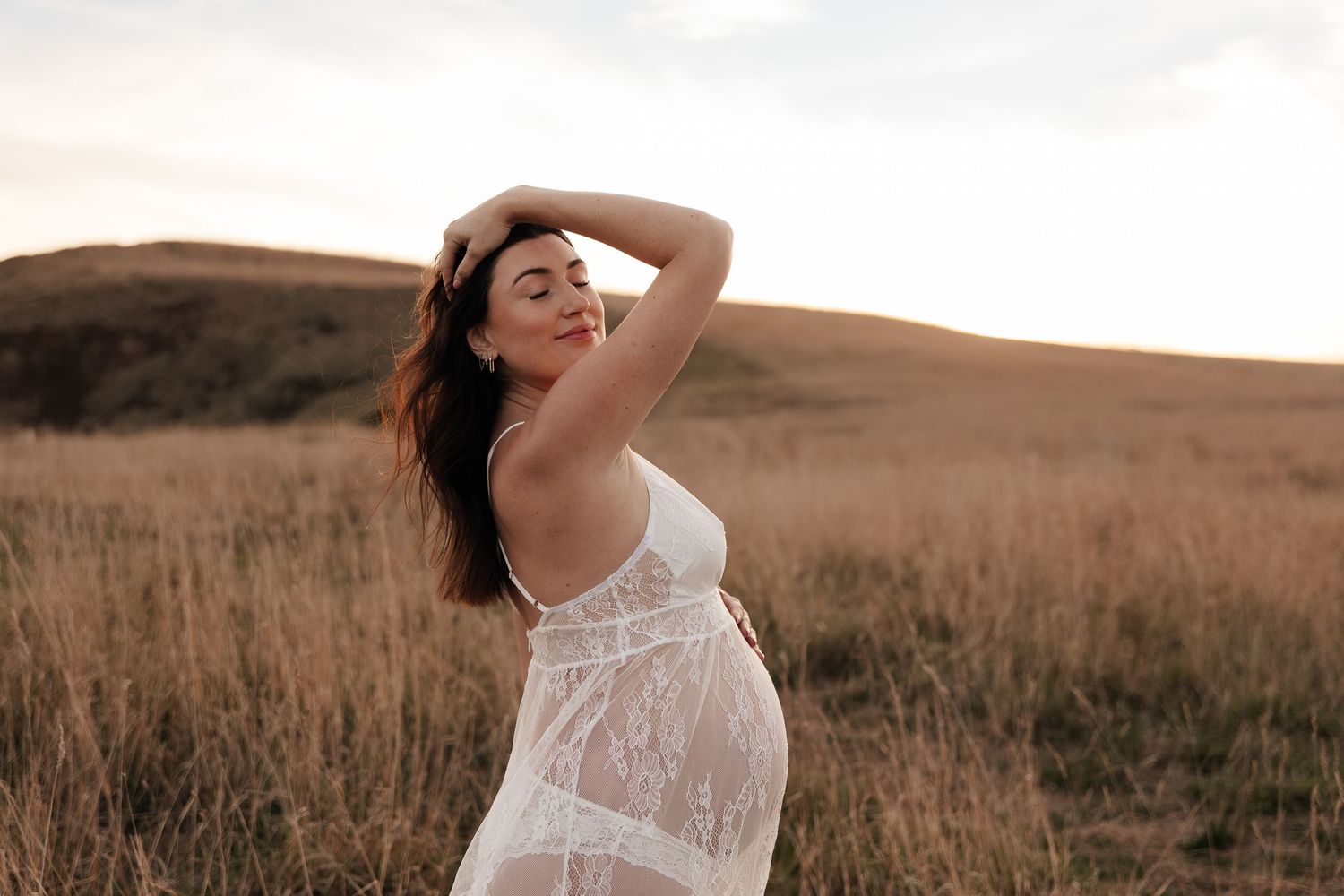 Silhouette of pregnant figure in white dress against ocean backdrop at sunset.