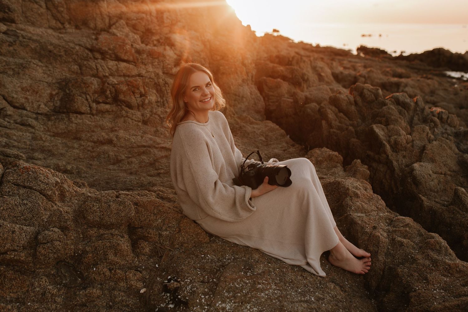 Woman in flowing white dress poses on coastal rocks during sunset.