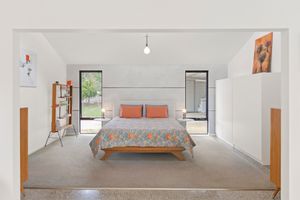 Bright minimalist bedroom with orange accent pillows, a colorful quilt, wooden bed frame, and large windows with garden views.