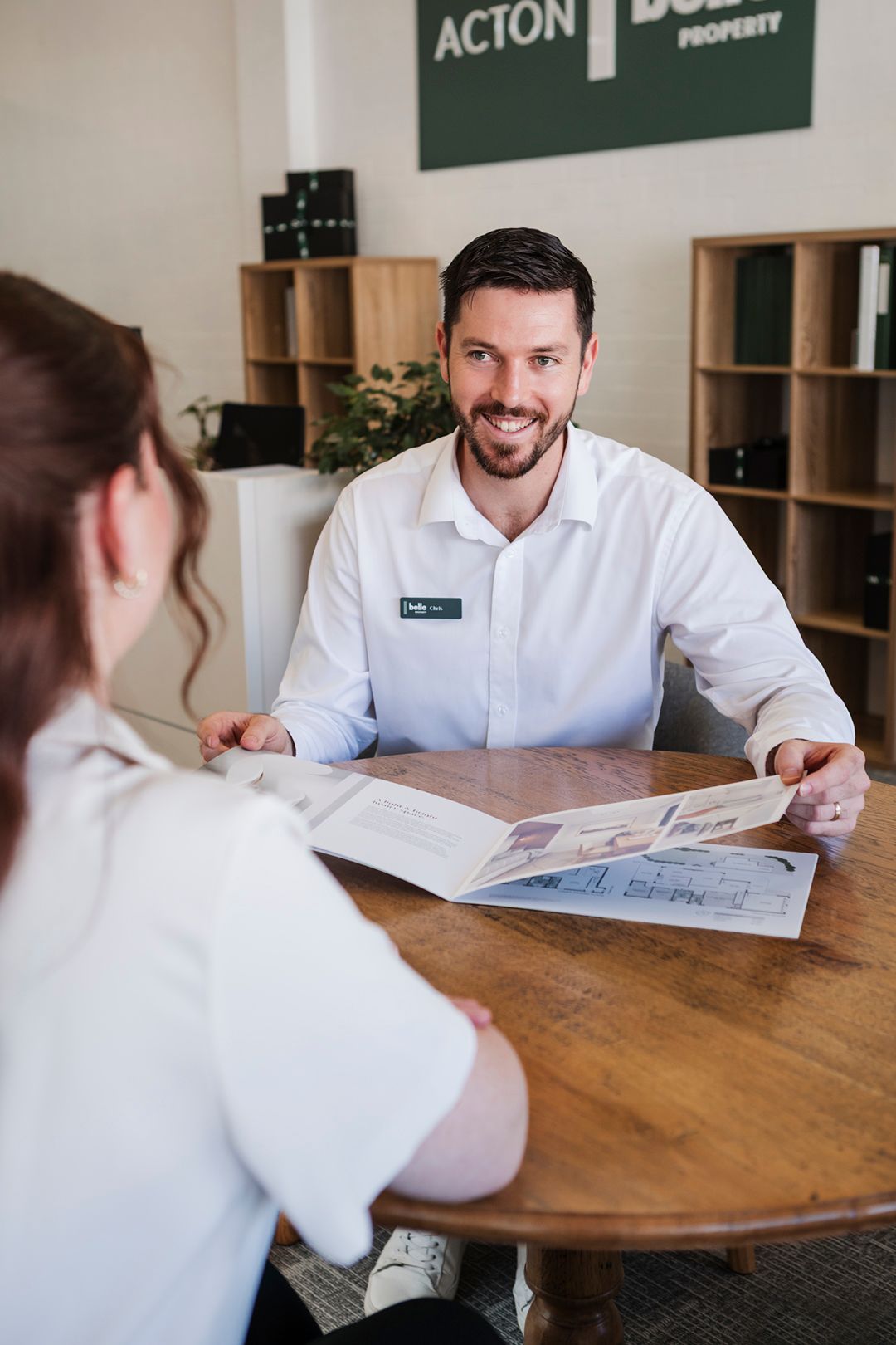 A smiling real estate agent discusses property brochure with client at wooden table in modern Acton Property office.