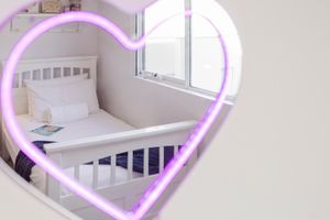 Heart-shaped mirror with pink neon light reflecting a white baby crib in a bright nursery room.