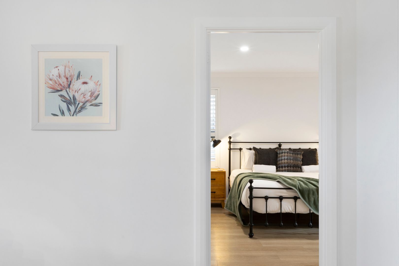 Hallway leading to a cozy bedroom with a black metal bed frame, green bedding, and a floral artwork on the white wall.