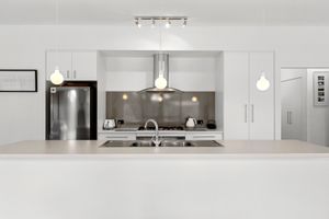 Modern white kitchen with stainless steel sink, range hood, and pendant lights over a sleek white countertop.