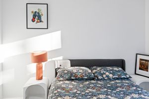 Cozy bedroom with floral bedding, dark headboard, orange lamp on white nightstand, and framed art on light gray walls.