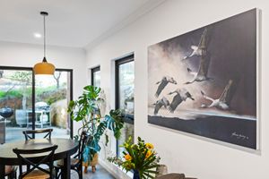 Modern dining area with monstera plant, pendant light, and large bird painting on white wall near glass doors.