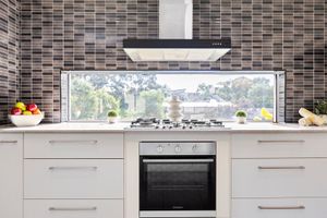 Modern white kitchen with mosaic tile backsplash, stainless steel range hood, gas cooktop, and oven with garden window view.