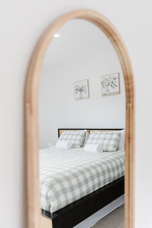 Arched wooden mirror reflecting a cozy bedroom with a gingham bedspread, white pillows, and floral wall art.