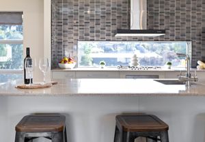 Modern kitchen island with white quartz countertop, bar stools, wine bottle, and gray mosaic tile backsplash.