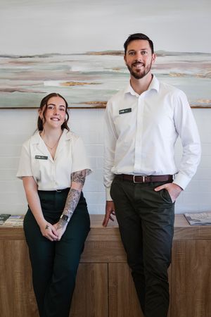 Two smiling hotel staff members in uniform pose together at a wooden reception desk with artwork behind them.