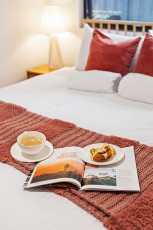 Cozy bedroom with white bedding, red pillows, open magazine, cup of tea, and pastry on a knit blanket.