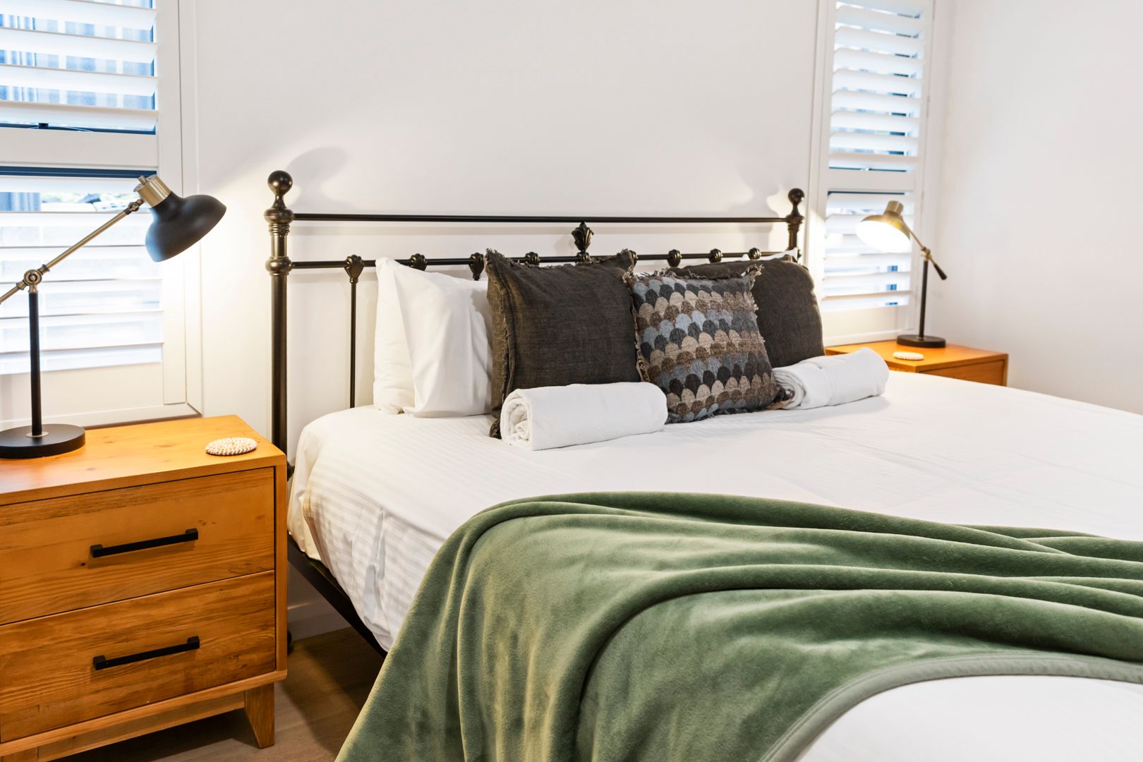 Cozy bedroom with iron bed frame, green throw blanket, white pillows, wooden nightstands, and bedside lamps.