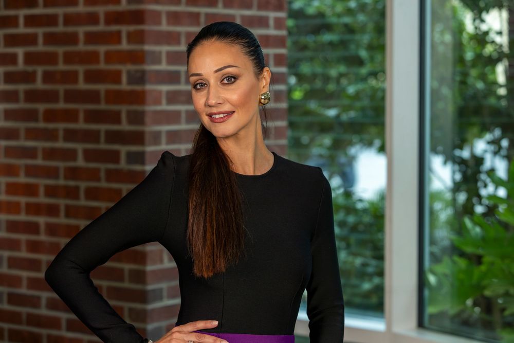 Smiling woman in black long-sleeve top and purple skirt poses confidently indoors near brick wall.