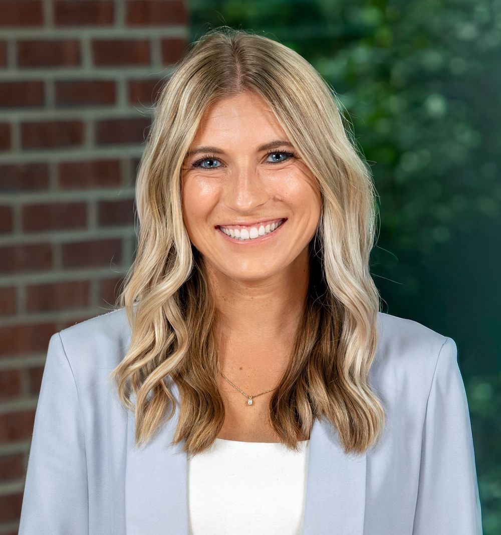 Smiling young professional woman with blonde hair wearing a light blue blazer and white top outdoors.