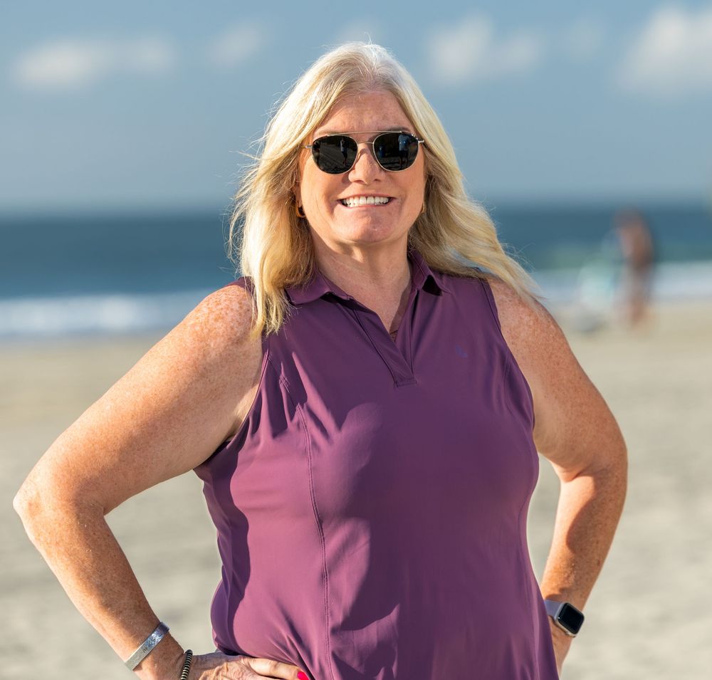 Smiling blonde woman in purple sleeveless top posing on sunny beach with ocean waves in background.