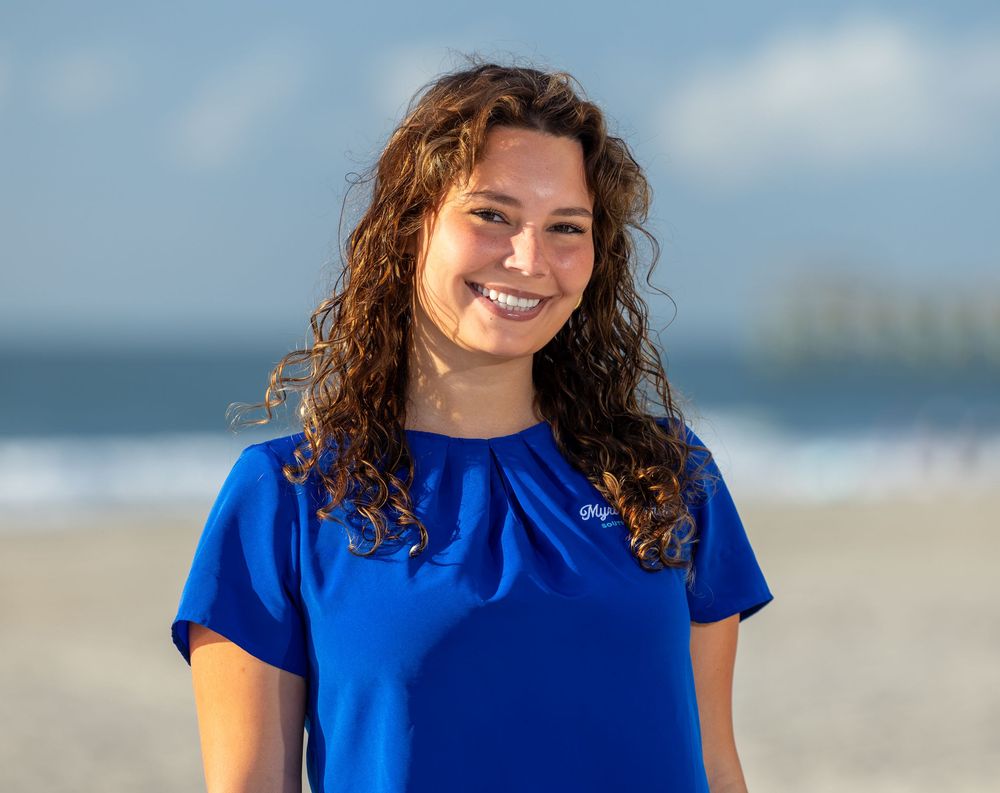Smiling woman with curly hair wearing a blue blouse on a sunny beach with ocean in the background.