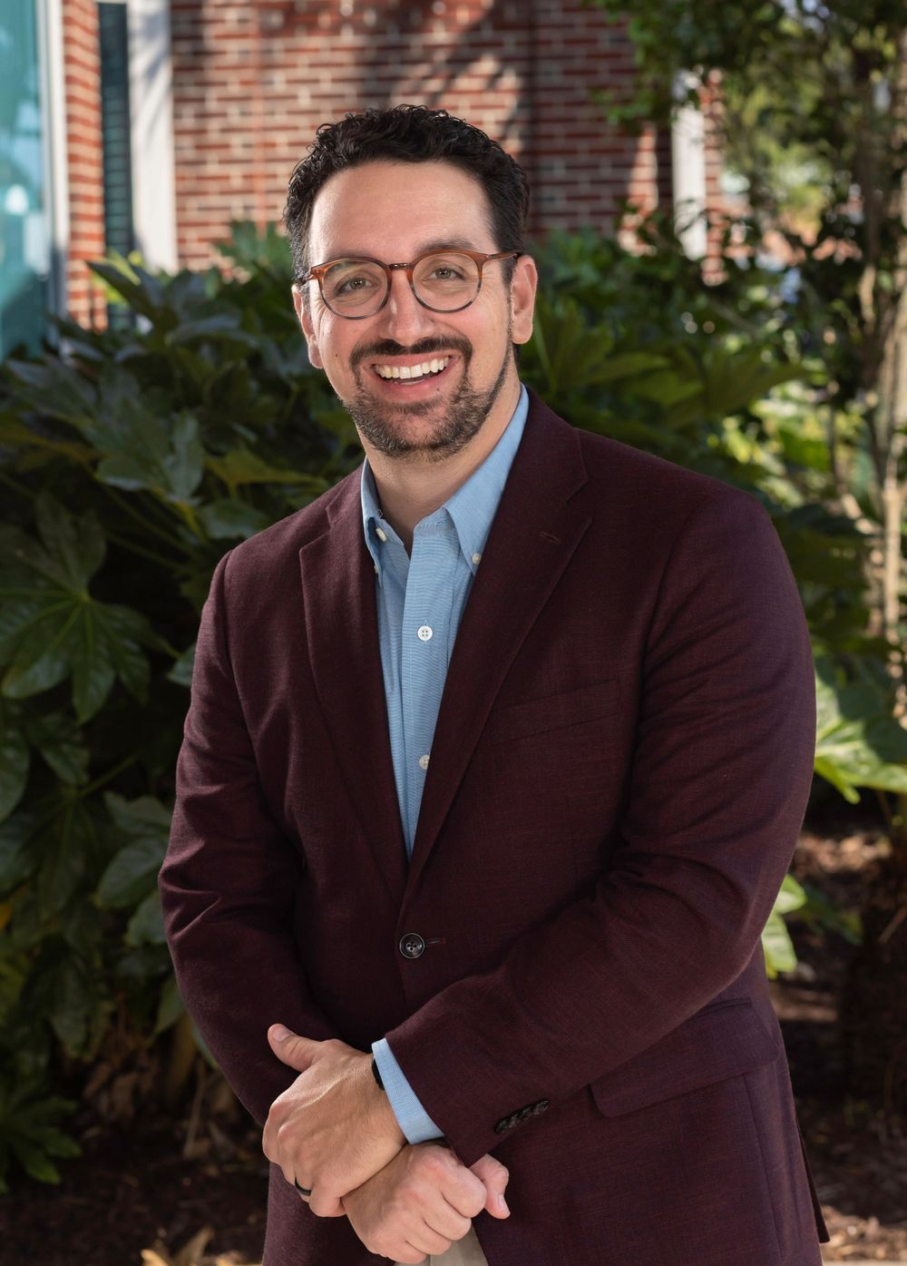 Smiling man in a maroon blazer and glasses poses outdoors among tropical greenery.