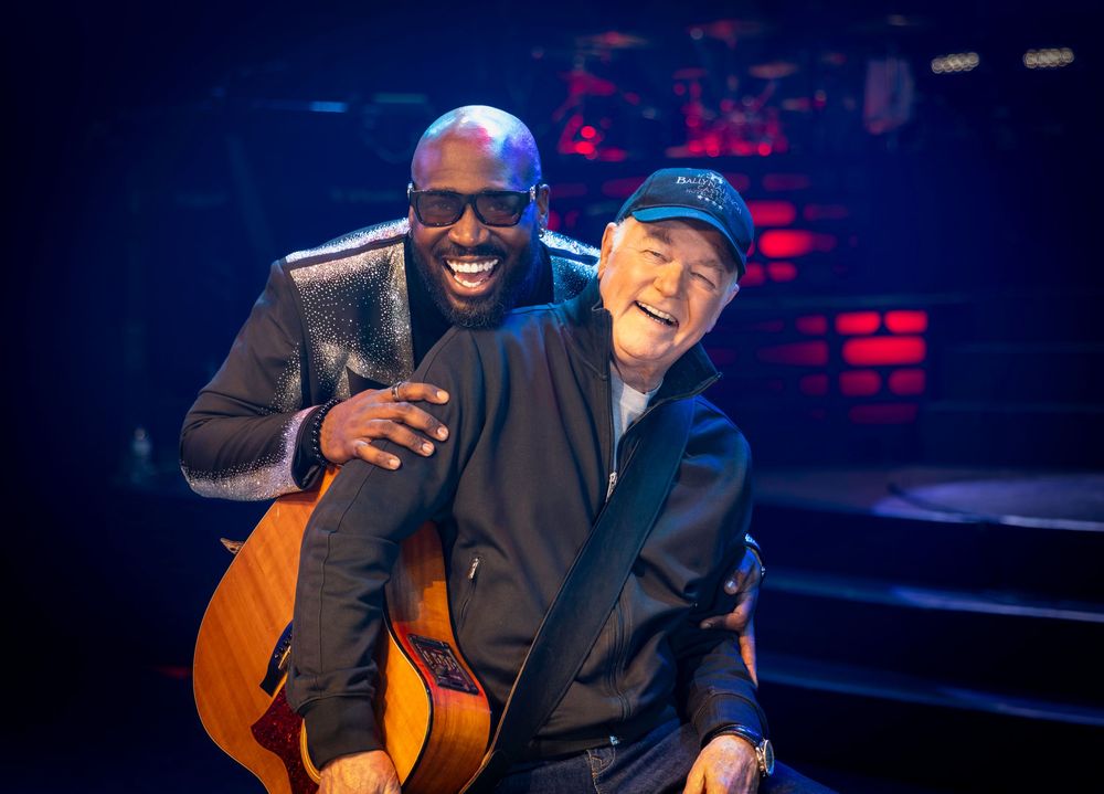 Two men smiling on stage, one holding an acoustic guitar under blue stage lighting.