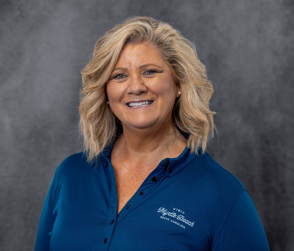 Professional headshot of a smiling blonde woman wearing a blue polo shirt against a gray background.