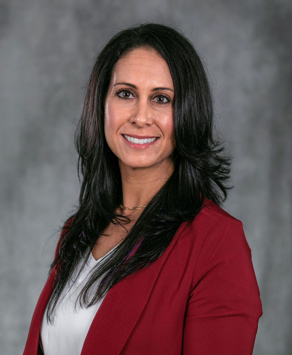 Professional headshot of a woman with dark hair wearing a red blazer, smiling confidently against a gray background.