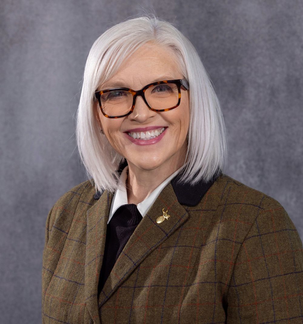 Professional woman with white hair and glasses wearing a plaid blazer smiling for a formal portrait photo.