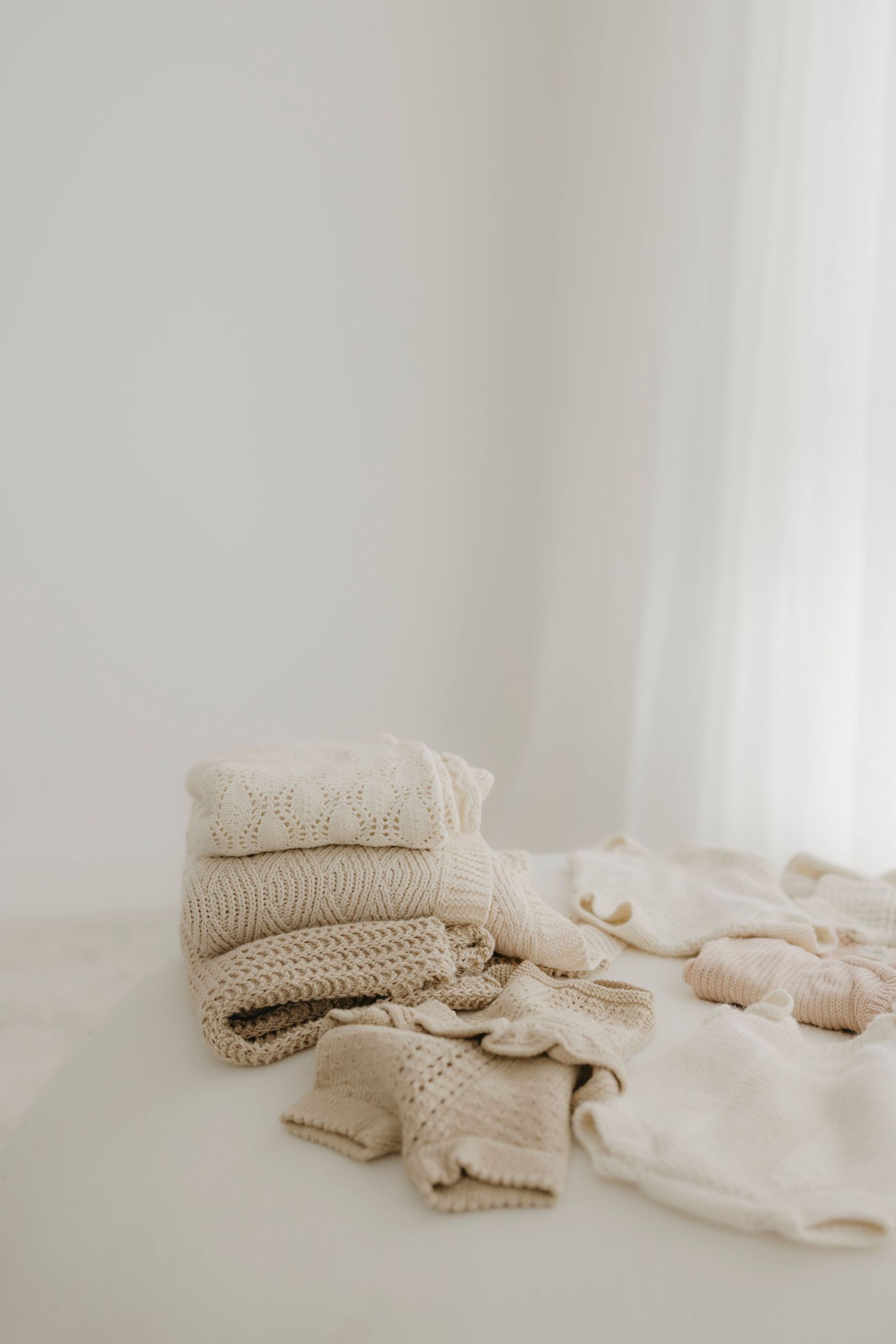 Stacked cream knit blankets and soft baby garments arranged on a white floor near sheer curtains.
