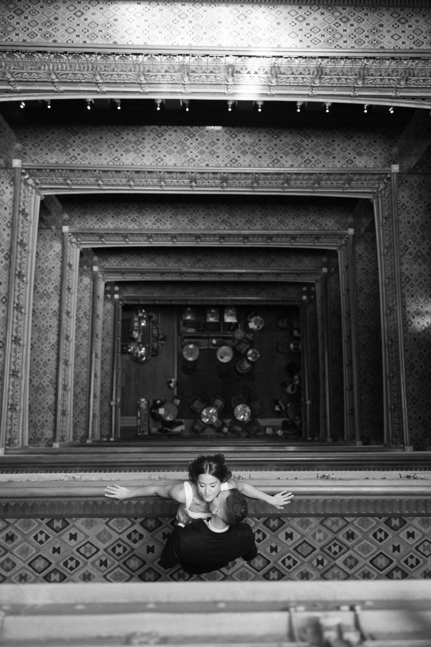 Black and white aerial view of a couple sharing a kiss on an ornate decorative balcony ledge.