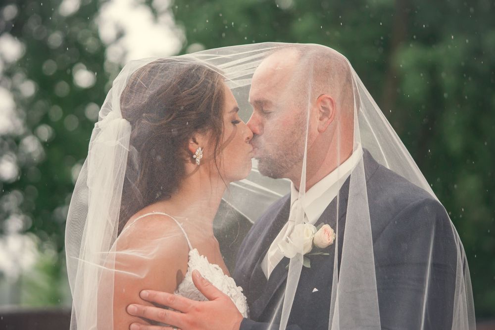 A romantic wedding couple shares tender kisses under a flowing white veil in a garden setting.
