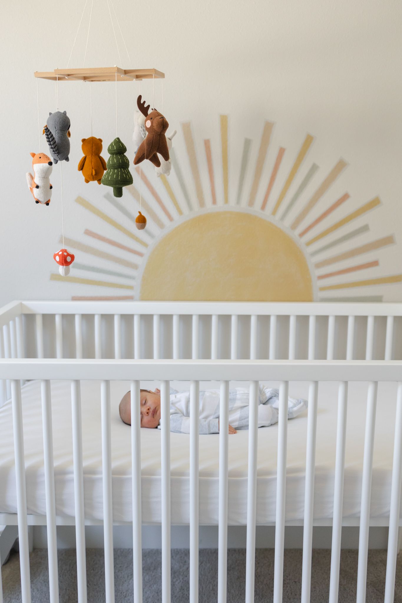 Nursery room with white crib and woodland animal mobile hanging above, featuring a painted sun design on the wall.