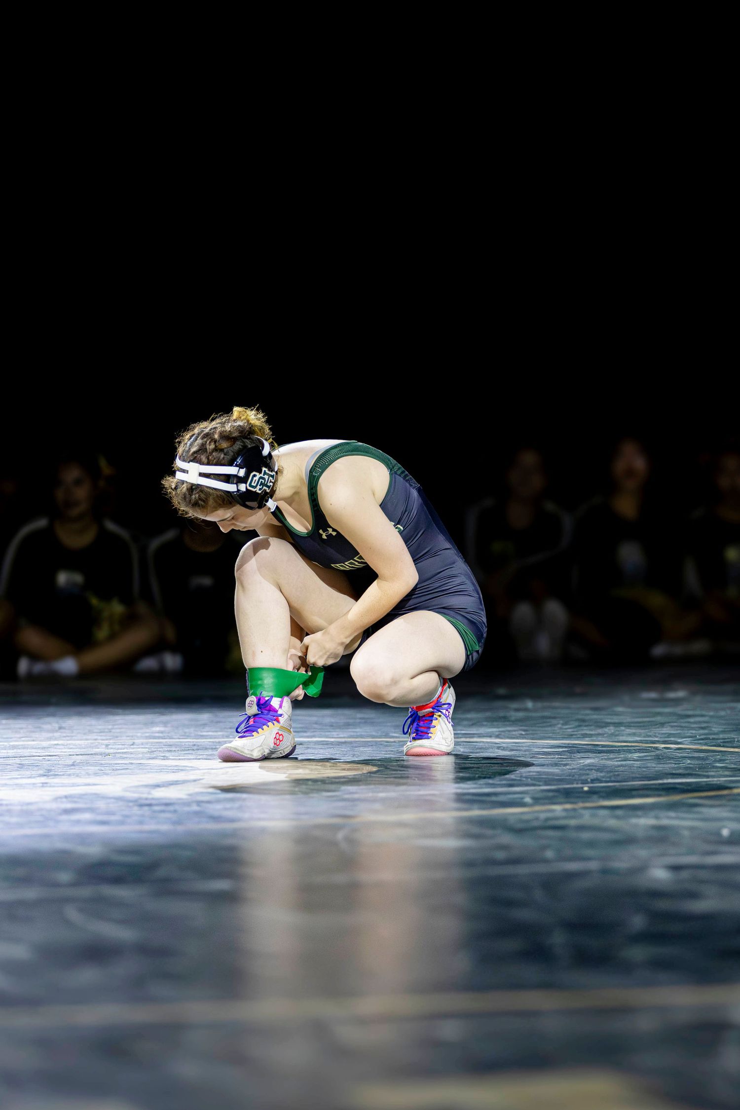 Female wrestler in green and navy uniform crouches in ready position on mat under spotlight during competition.