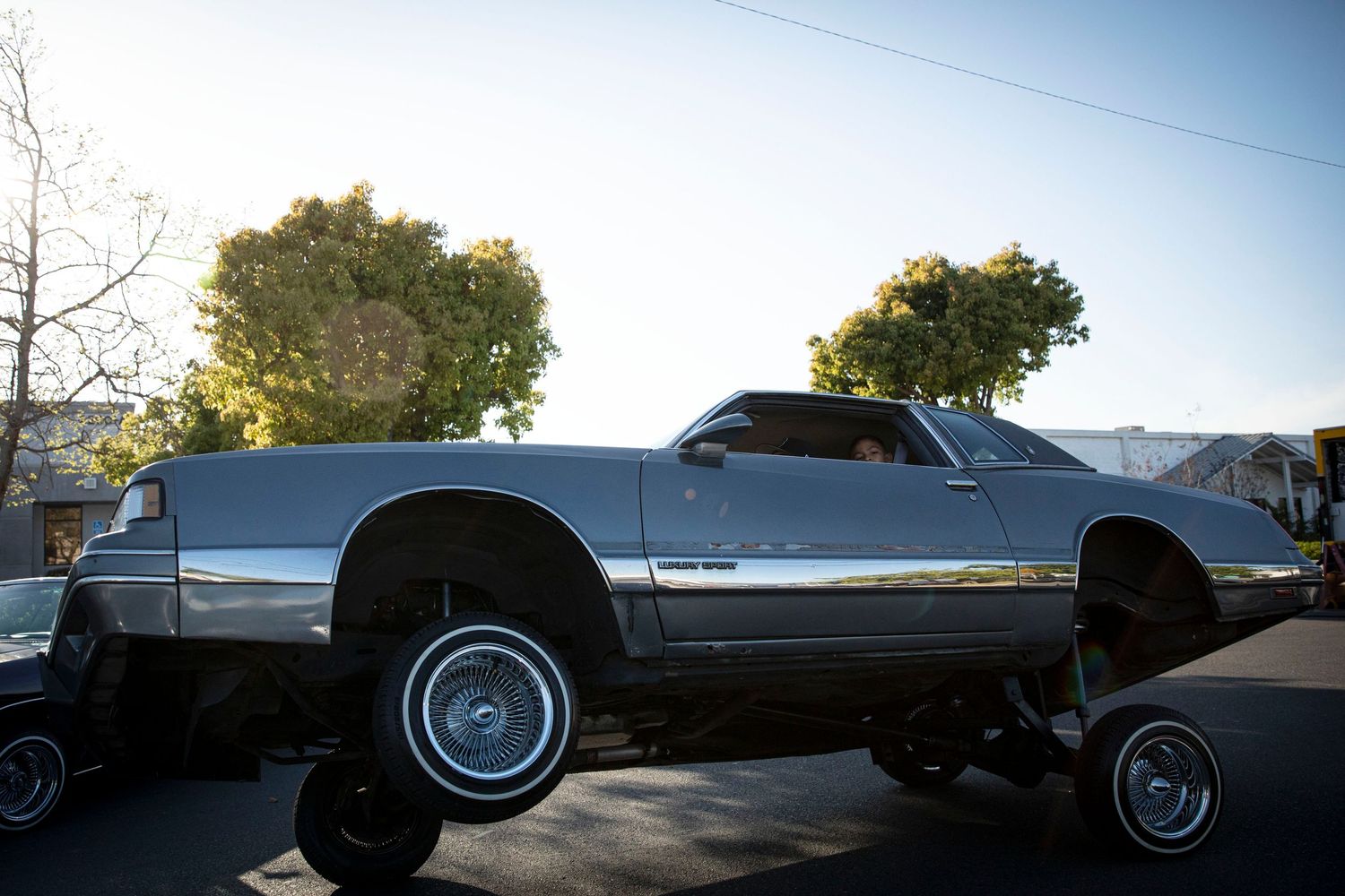 A silver lowrider car with hydraulics raised on one side, parked on a street with trees in the background.