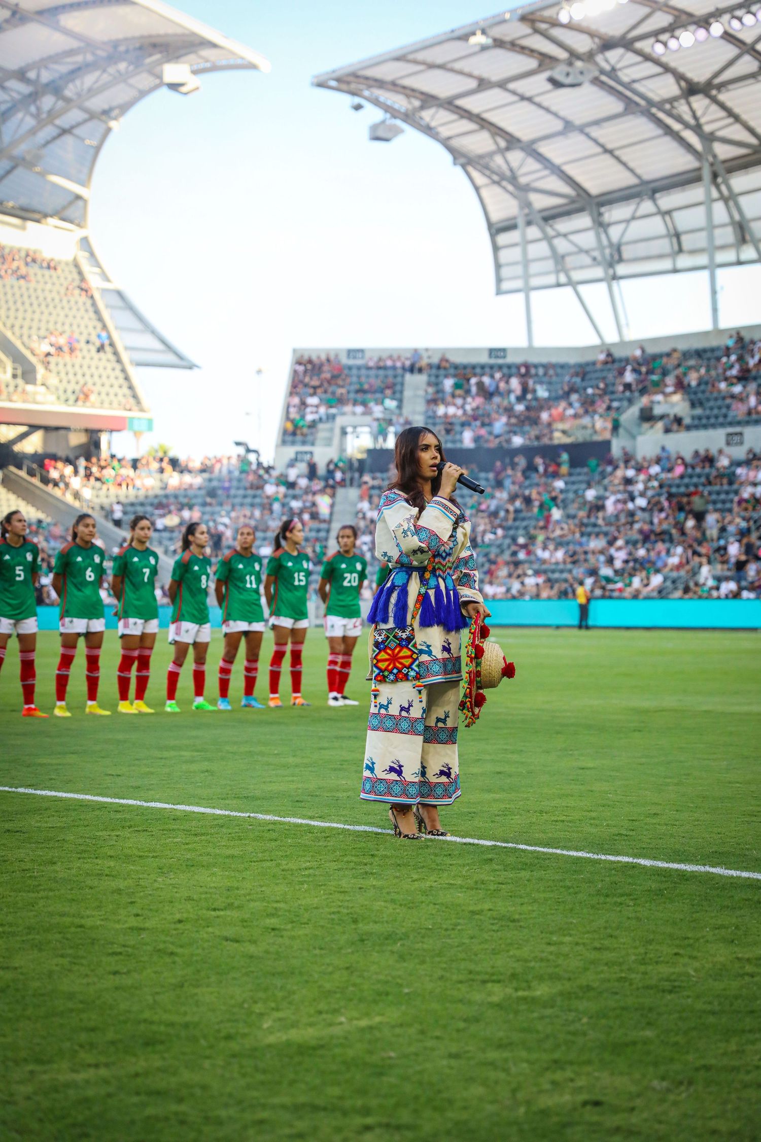 Woman in traditional Indigenous dress sings national anthem on soccer field before Mexico women's national team match.