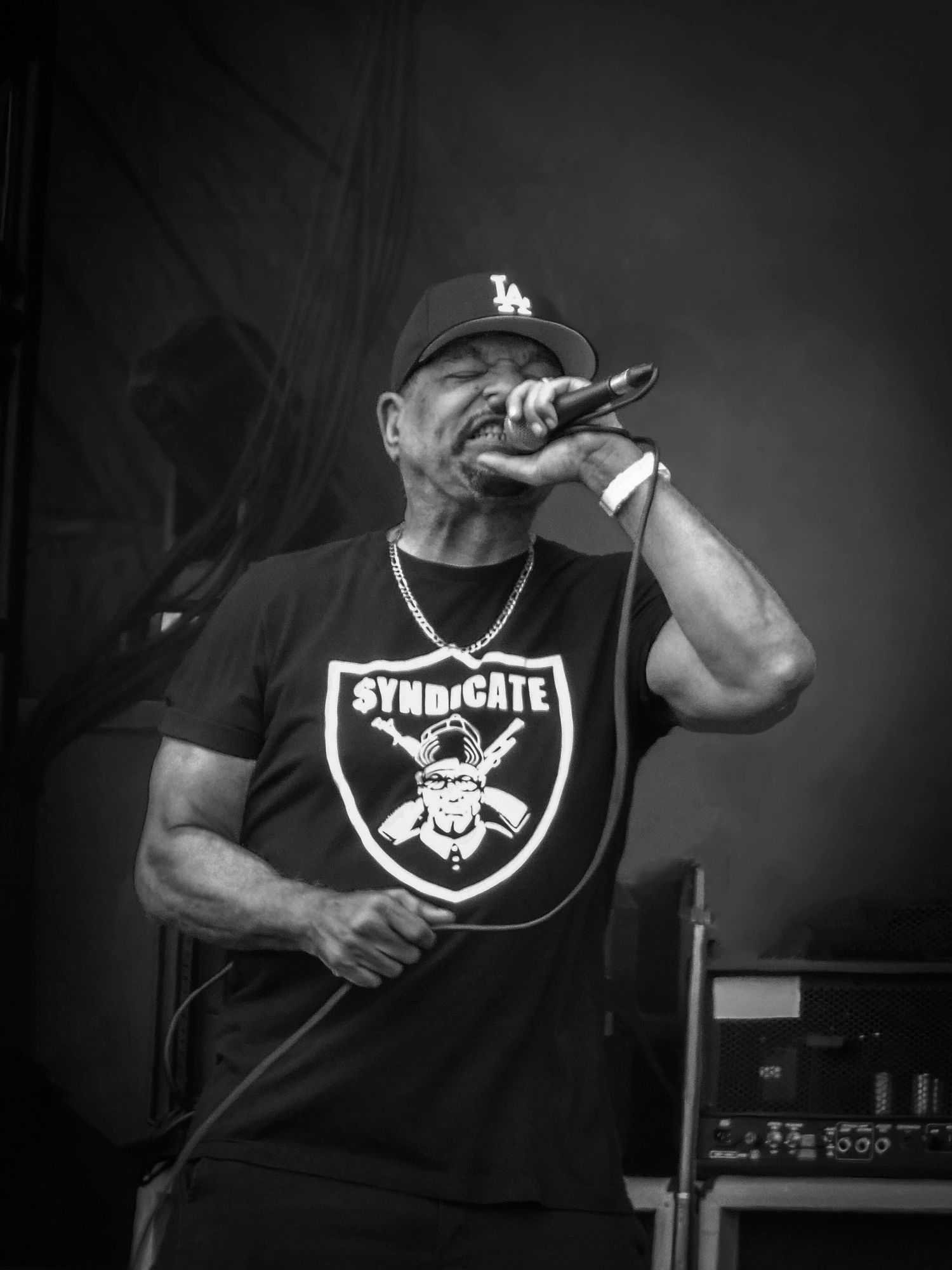 Black and white photo of a male rapper performing on stage, wearing an LA cap and Syndicate t-shirt, singing into a microphone.