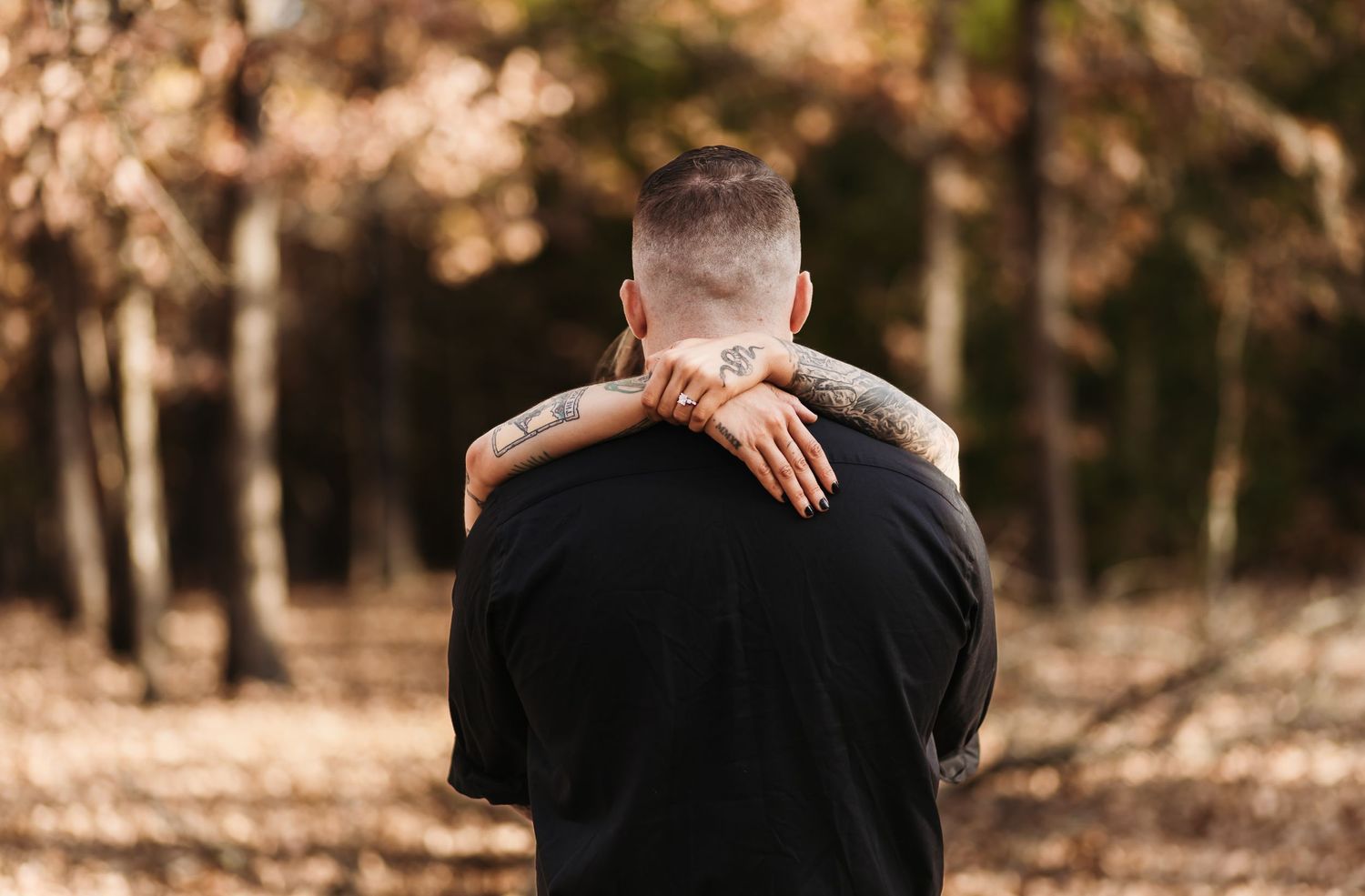 man's back facing the camera as the woman wraps her arms around his neck, highlighting her engagement ring