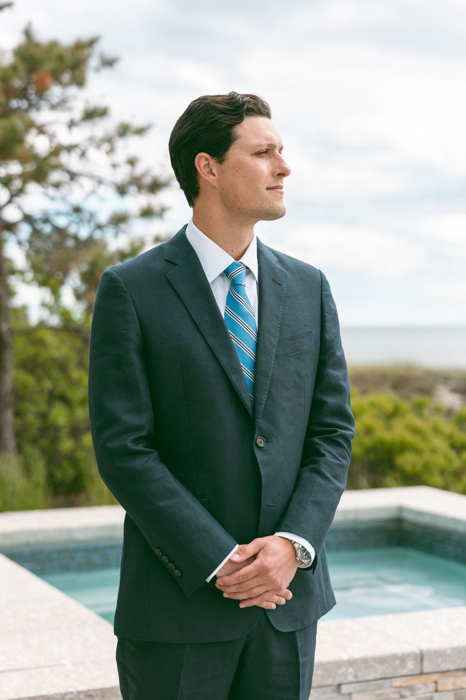 Handsome groom in dark navy suit and striped blue tie standing by outdoor pool with ocean view.