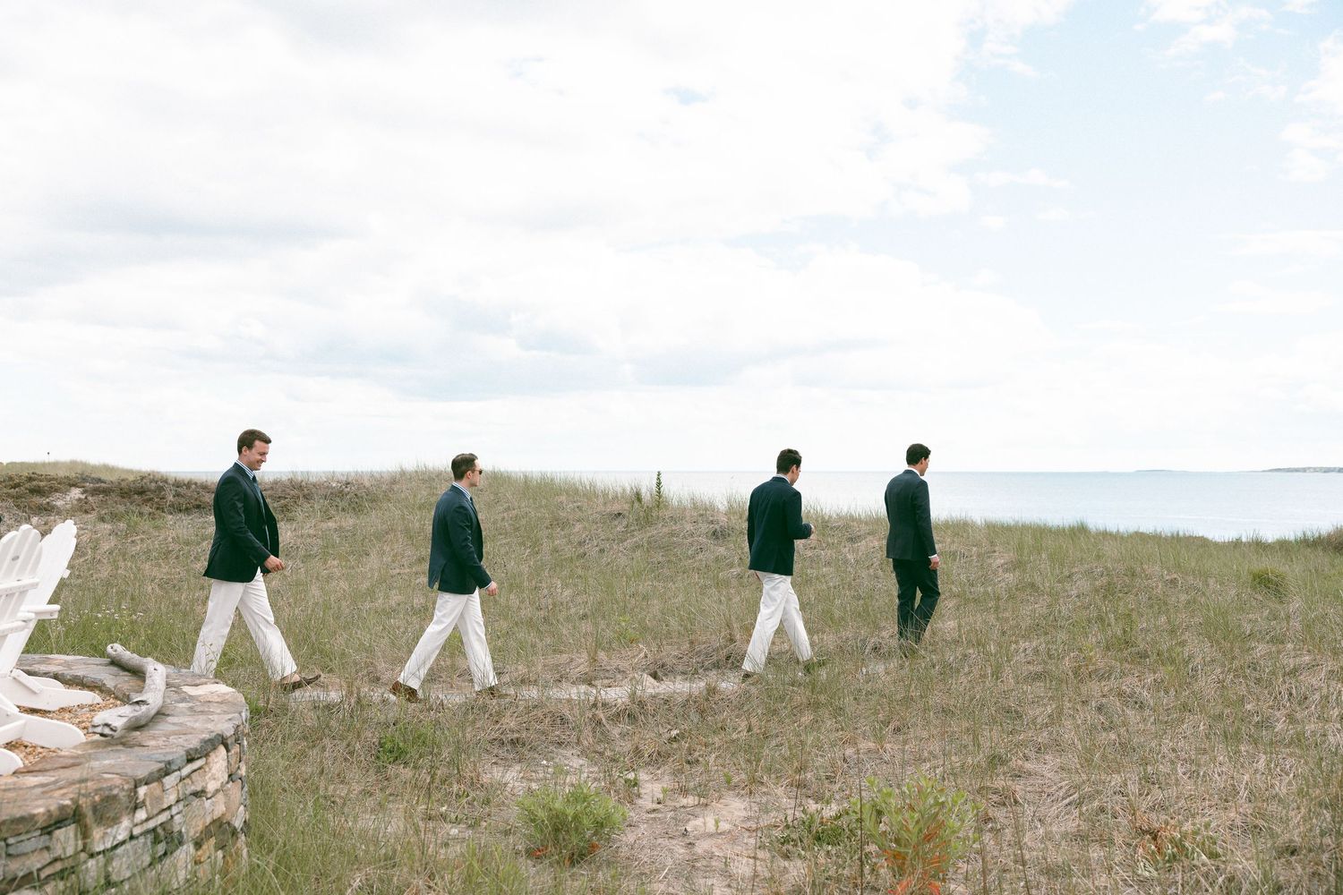 Four men in matching green blazers and white pants walking through coastal grasslands near the ocean.
