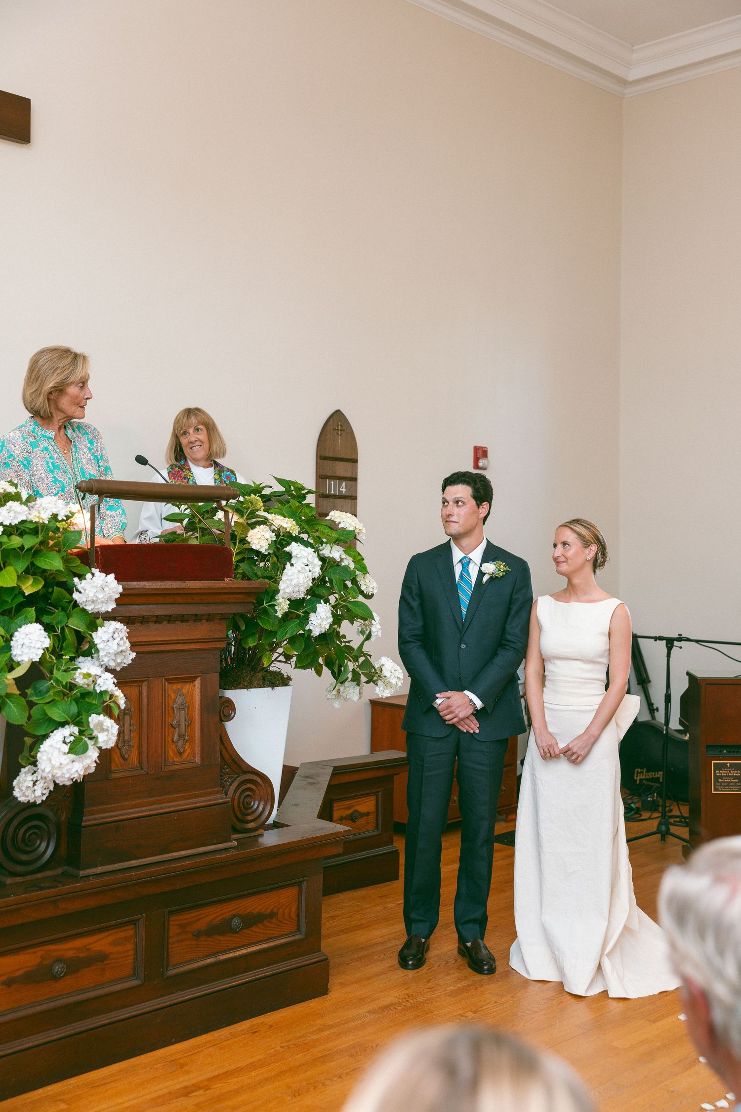 Bride and groom stand at altar during wedding ceremony while officiant speaks at wooden podium with white floral arrangements.