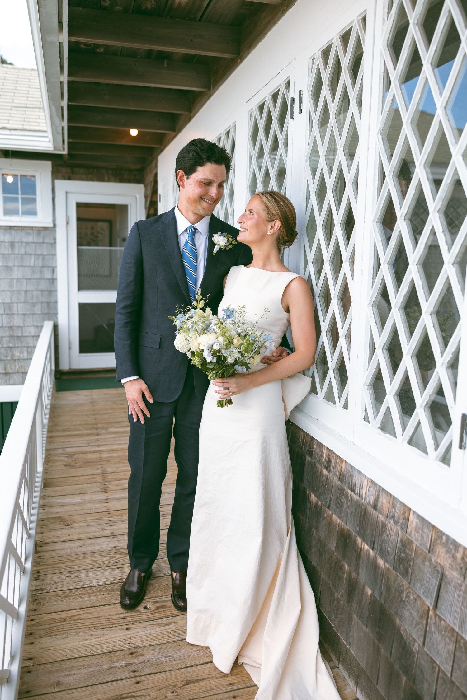 A bride and groom share a romantic moment on a wooden porch, she holds a white and blue bouquet.