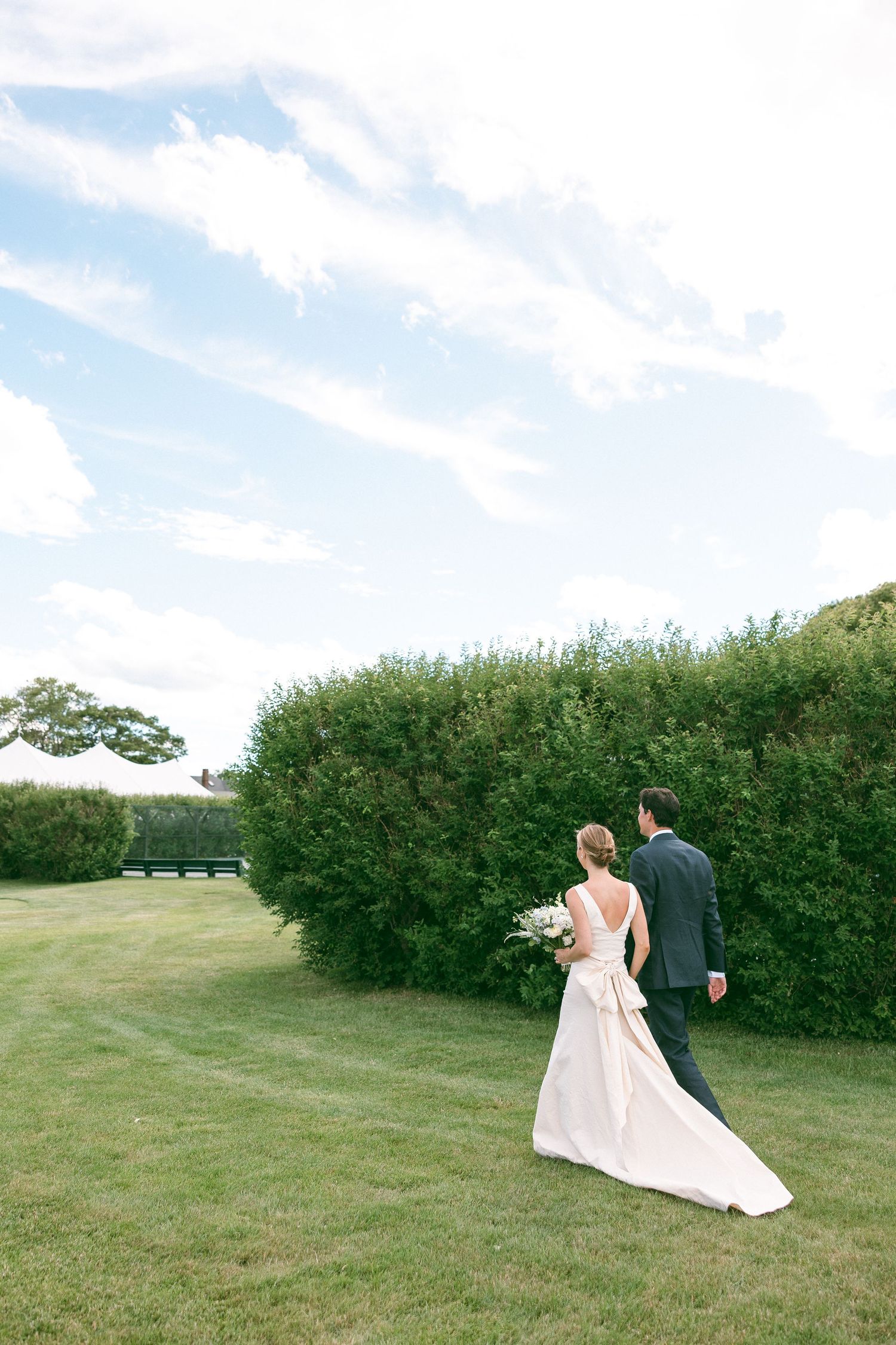 Bride and groom walk hand in hand past lush green hedges on manicured lawn under blue sky.