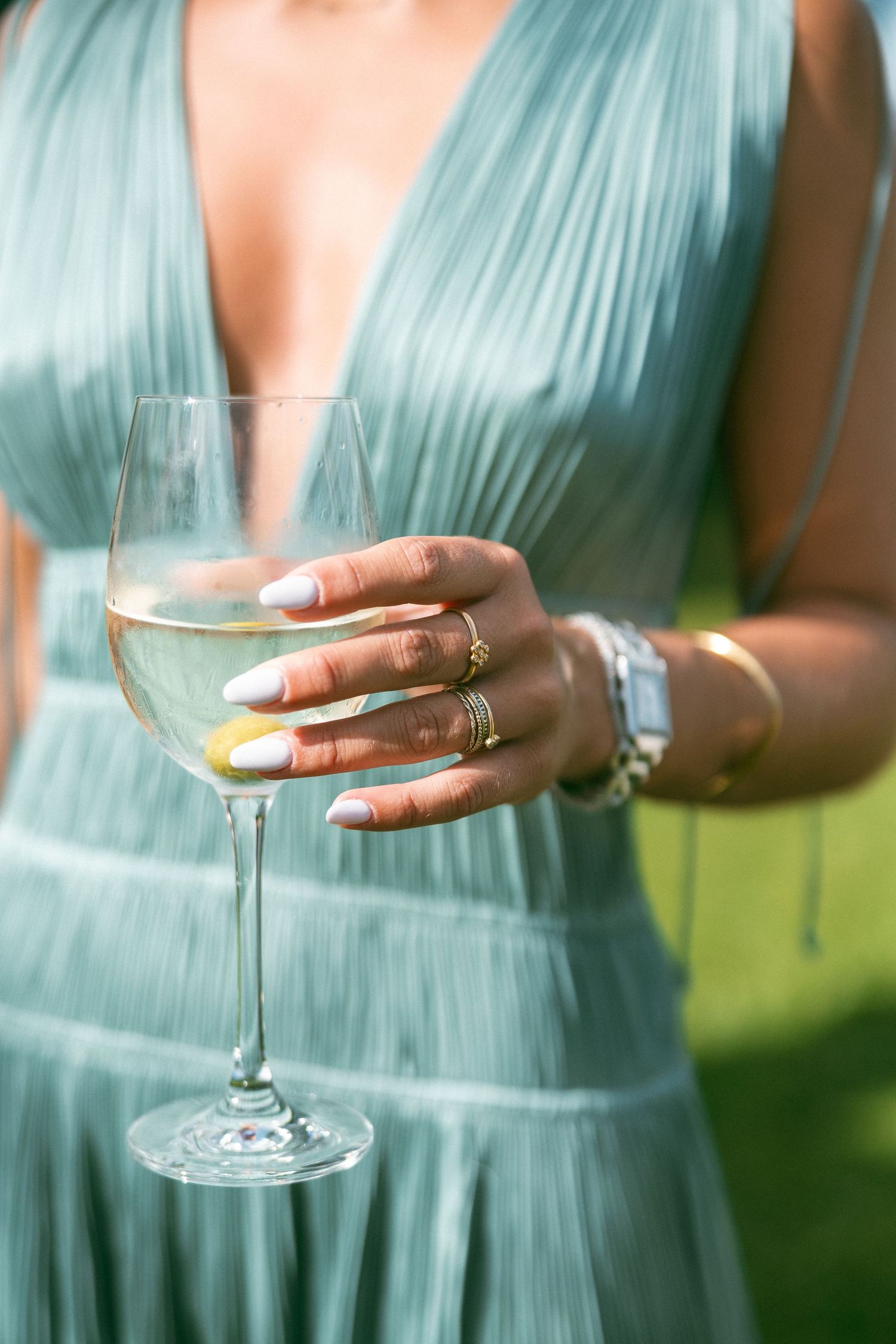 Woman in teal pleated dress holding a wine glass with white wine, adorned with gold rings and a bracelet.