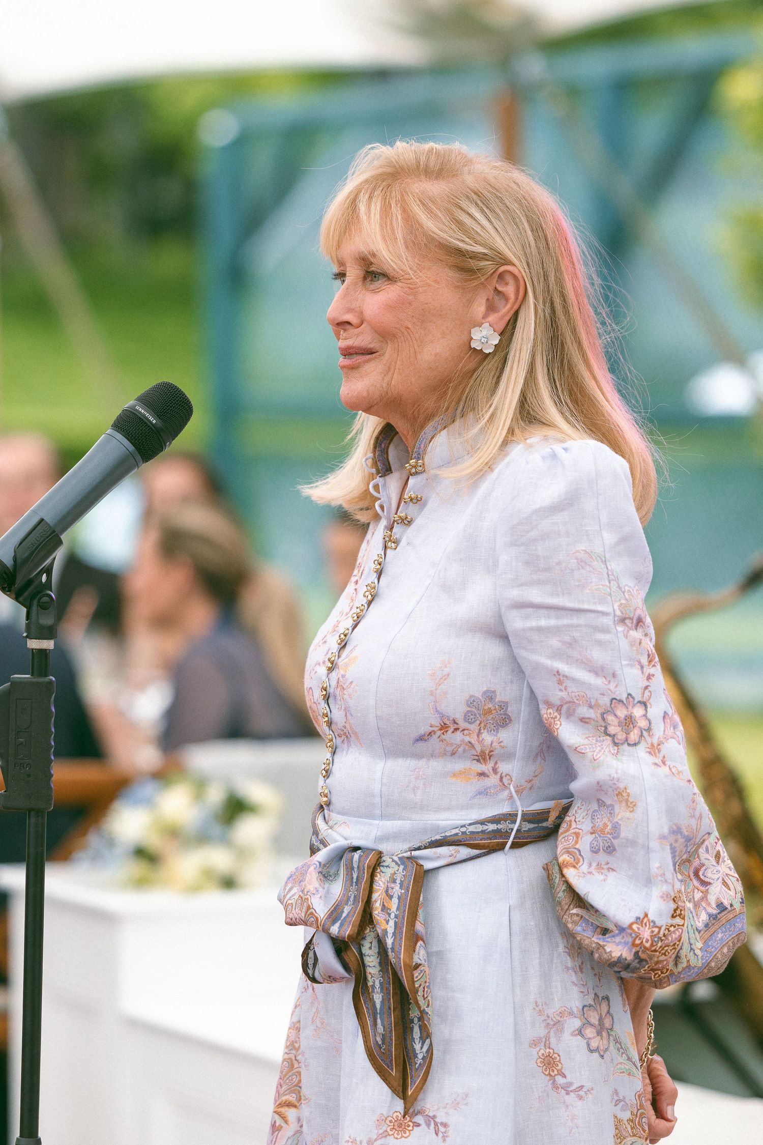 Elegant blonde woman in floral dress speaking at outdoor podium microphone at formal garden event.