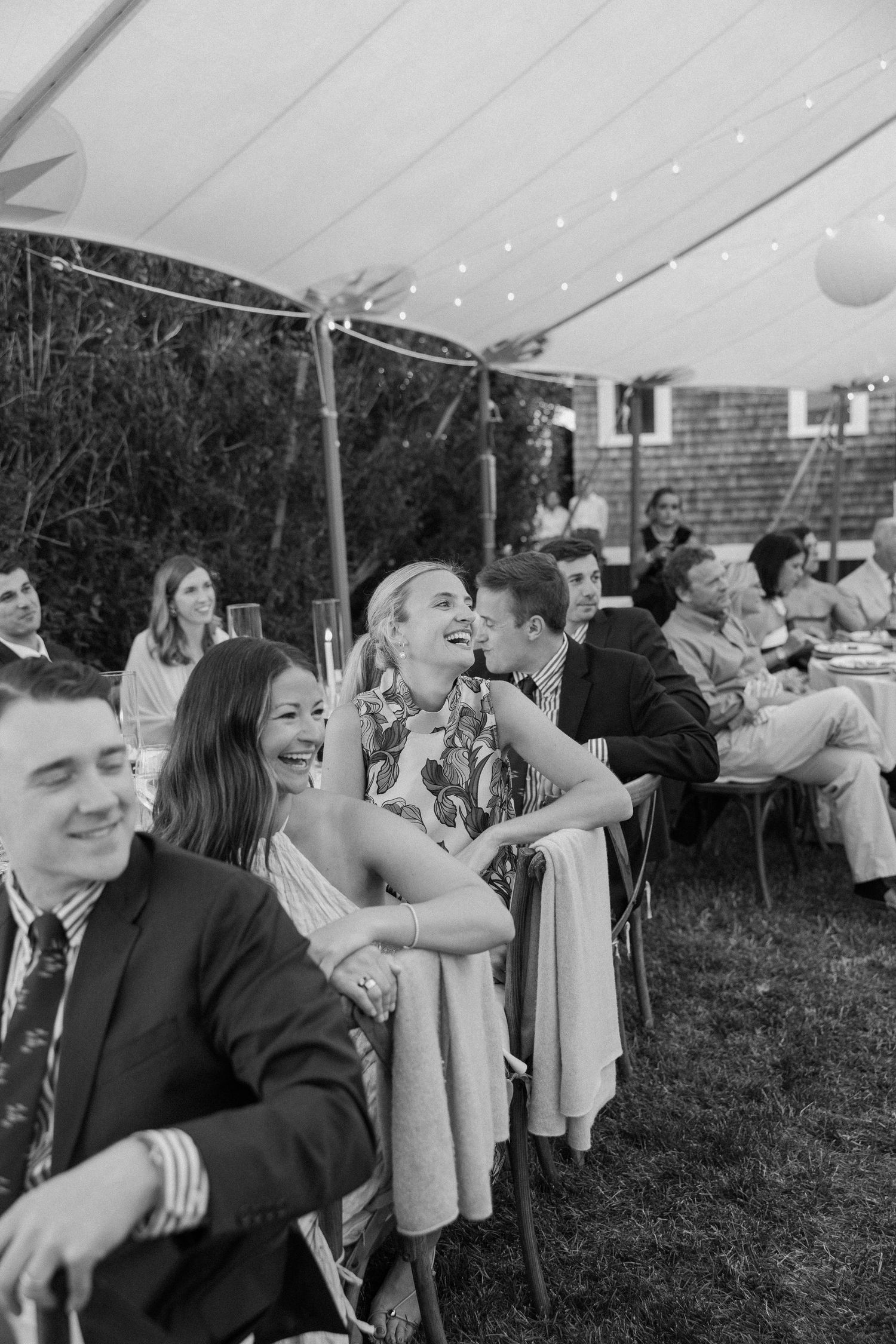 Wedding guests laughing and celebrating under a white tent with string lights at outdoor reception.