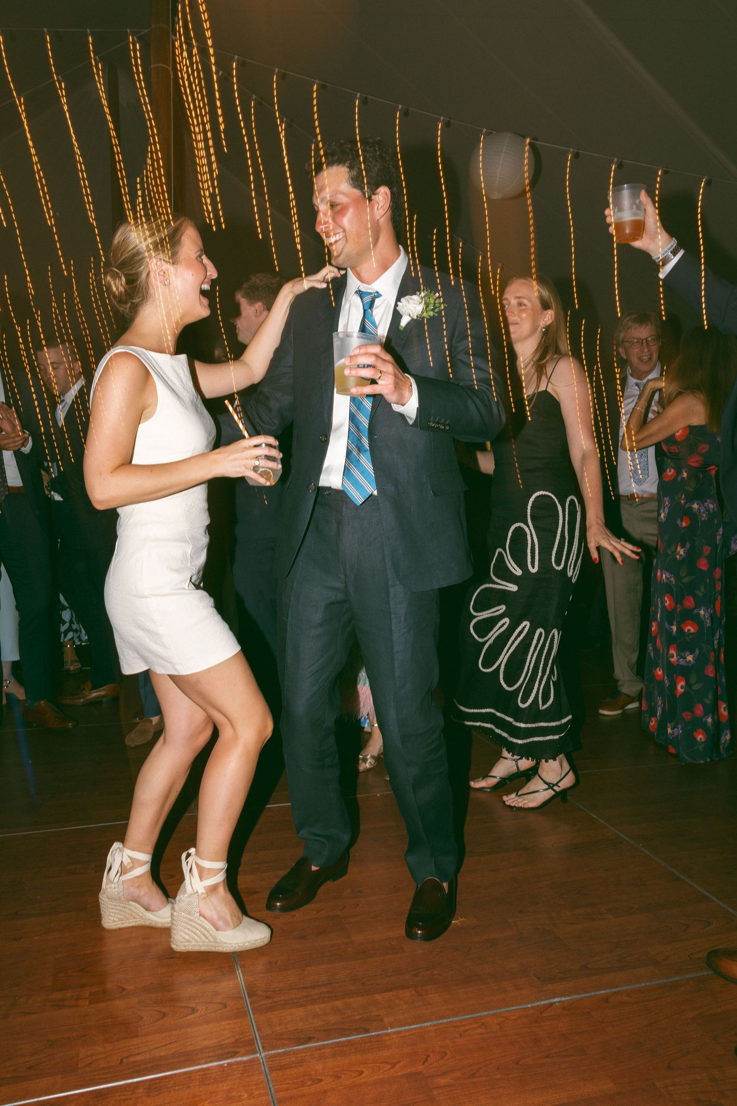 Couple dancing and laughing at a wedding reception in a tent with string lights.