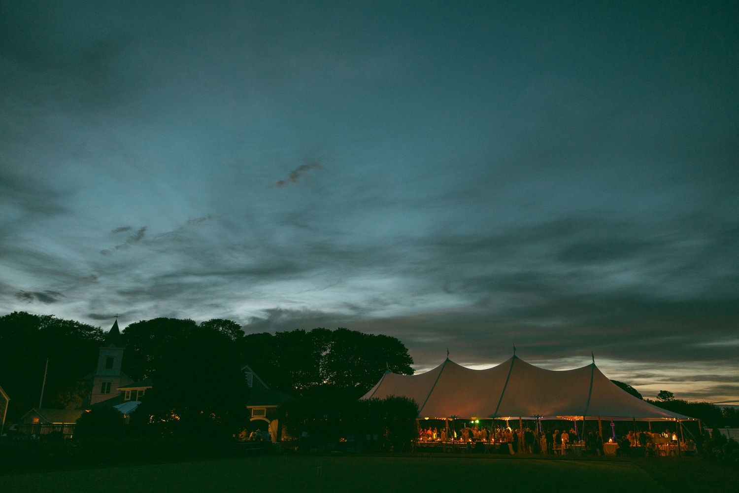 Illuminated white event tent glowing against a dramatic dusk sky surrounded by trees.