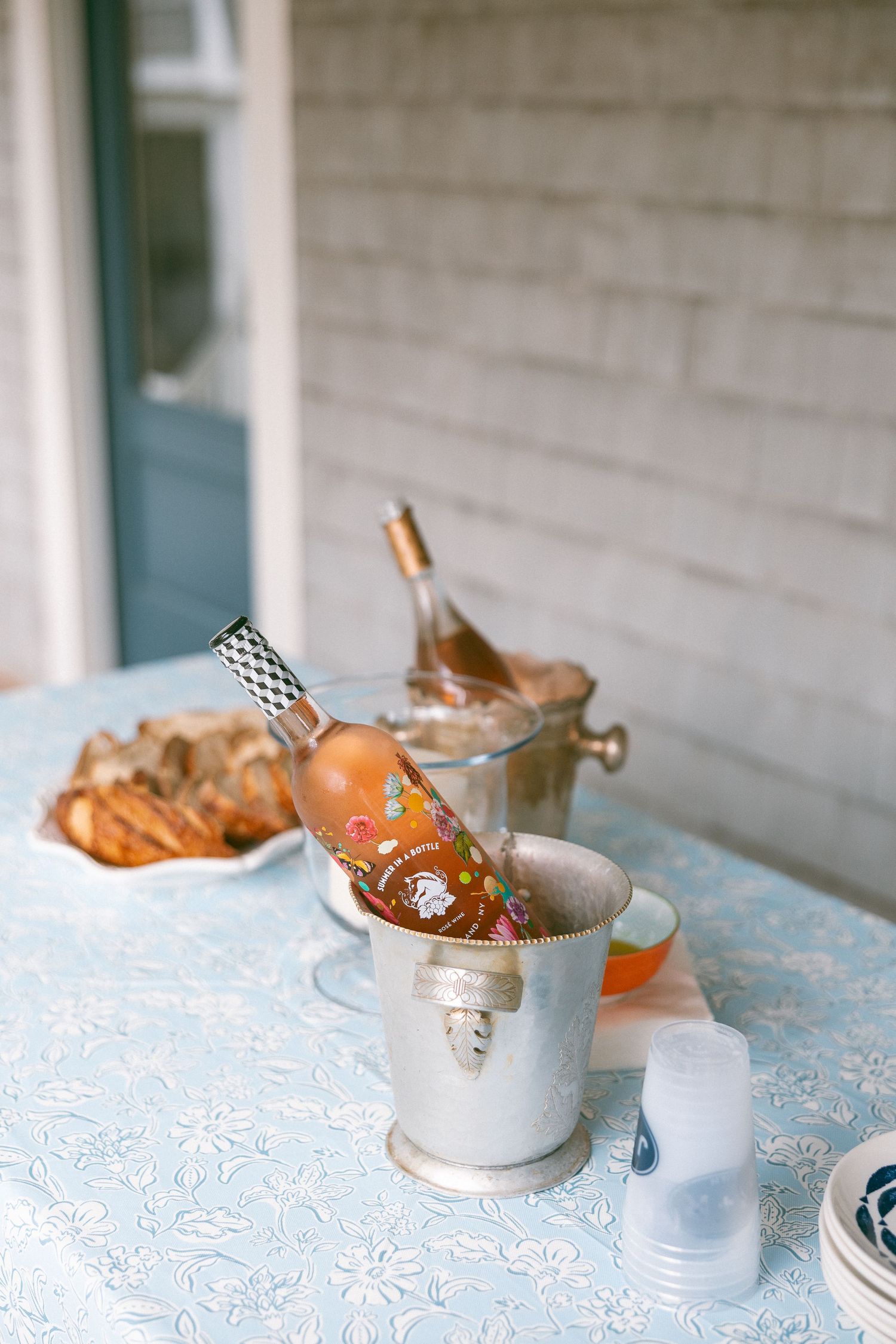 Rose wine bottle chilling in silver ice bucket on floral tablecloth at outdoor party with bread and glasses.