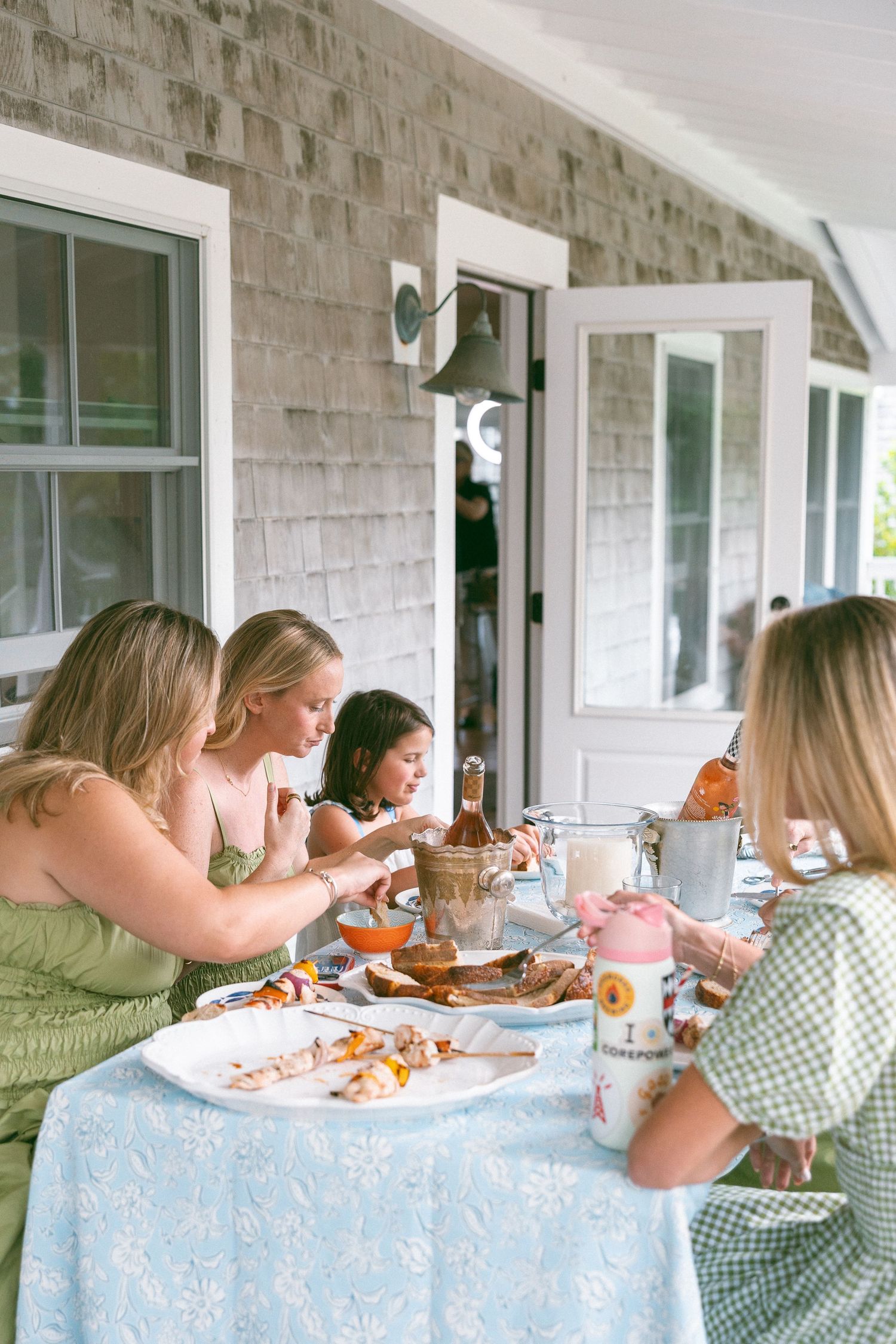 Women dining together on a porch of a shingle-style home, enjoying food on a floral blue tablecloth.