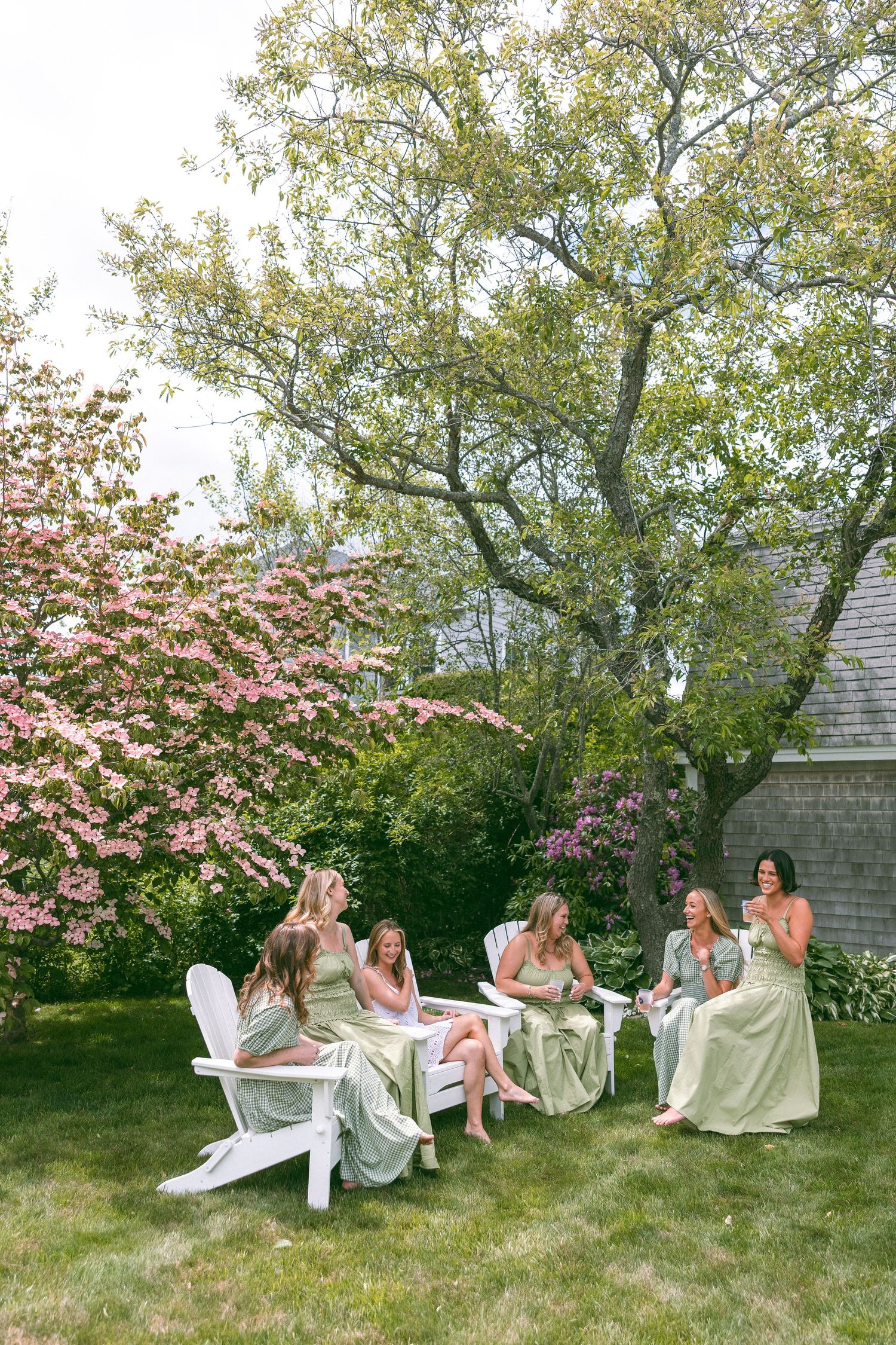 Bridesmaids in sage green dresses sit on white Adirondack chairs in a lush garden with blooming pink trees.