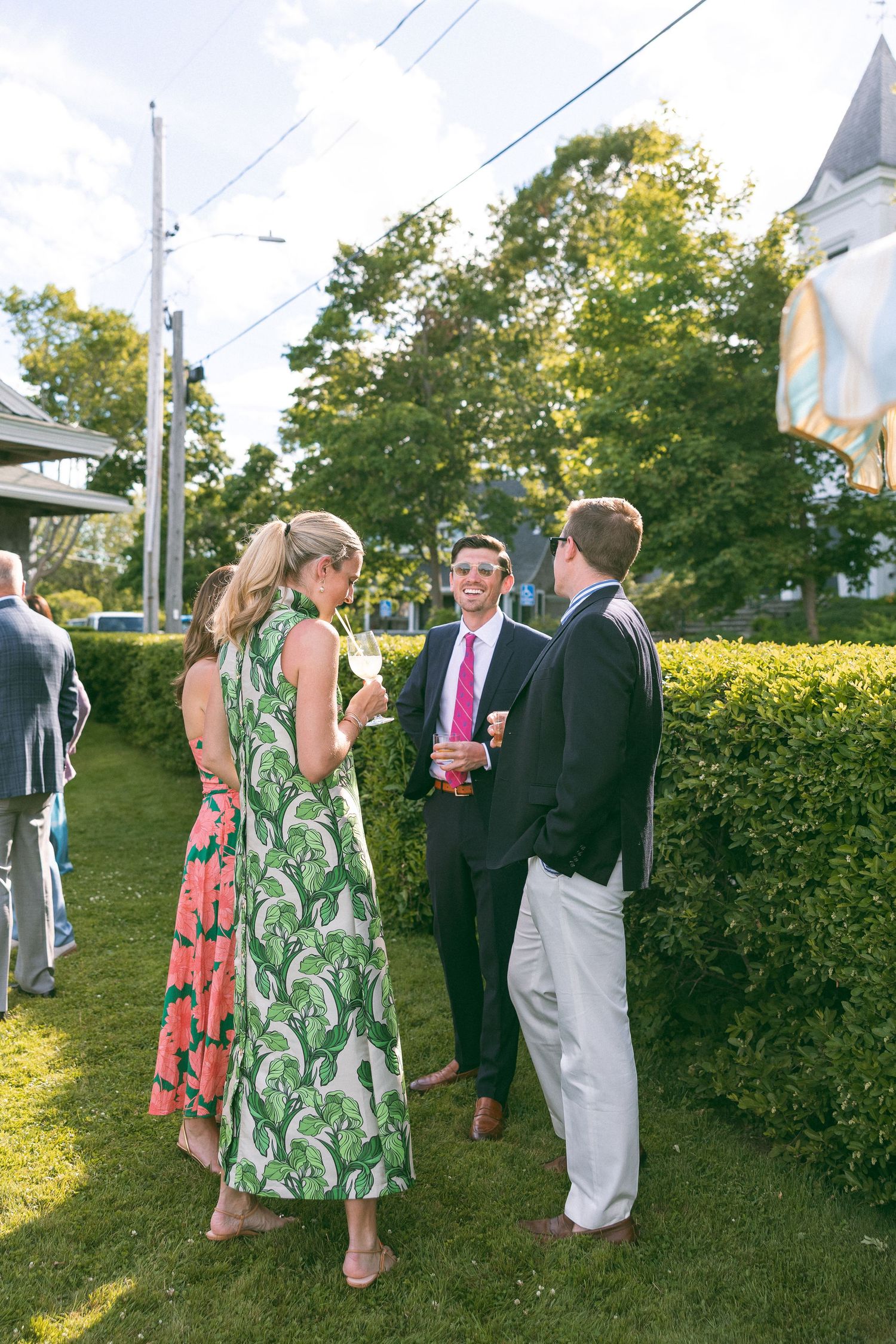 Guests in formal attire socialize outdoors on green lawn, woman in tropical print dress holds wine glass.