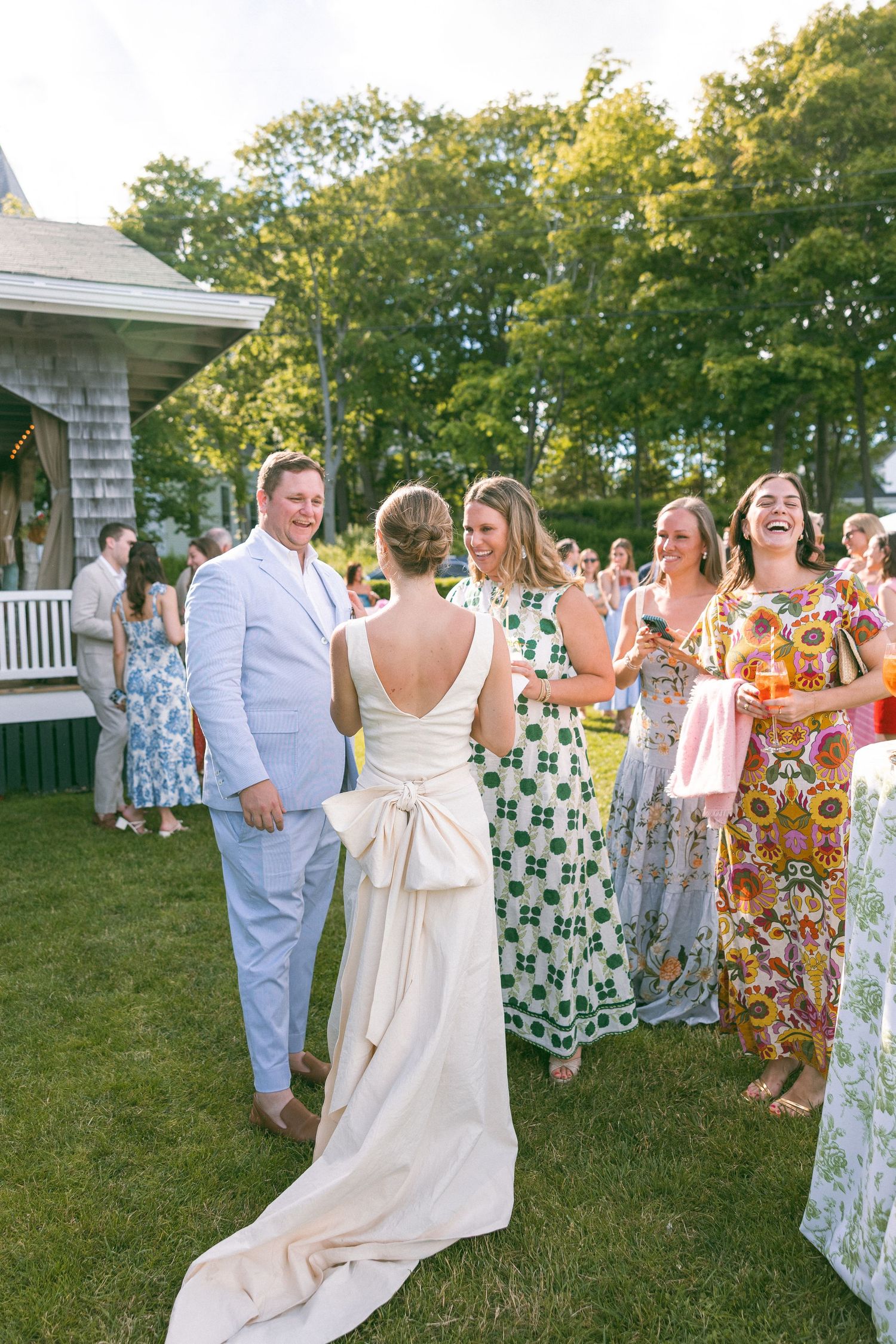 Bride in elegant backless white gown with bow greets smiling guests in colorful dresses at outdoor wedding reception.