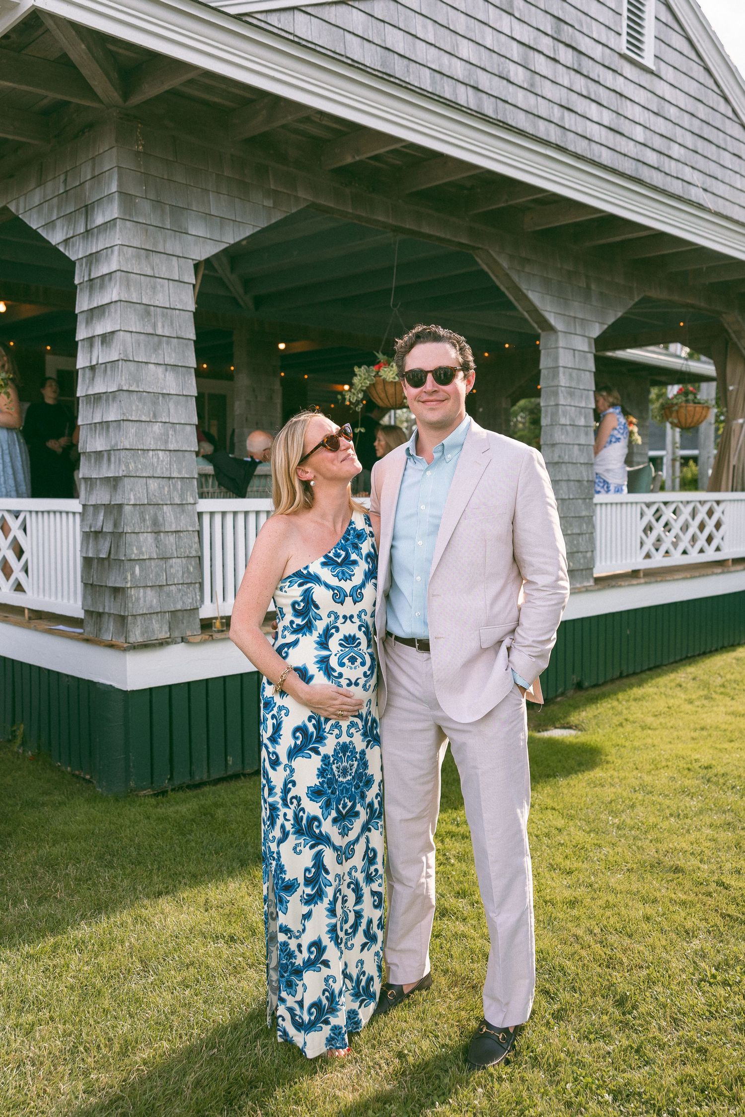 A couple poses outdoors at a summer event, woman in blue floral dress, man in tan suit, beside a shingle-style building.